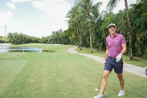 A golfer stands beside a tee box and water on the course.